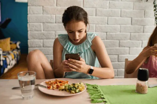 A teenage girl sits with a meal on a plate in front of her but is distracted and looking at her phone