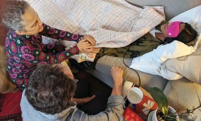 Two people oversee a blindfolded woman in bed