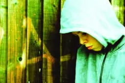 A young boy with a hood on stands next to a fence and looks dejected