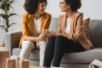 Two women sit on a couch talking to each other and smiling