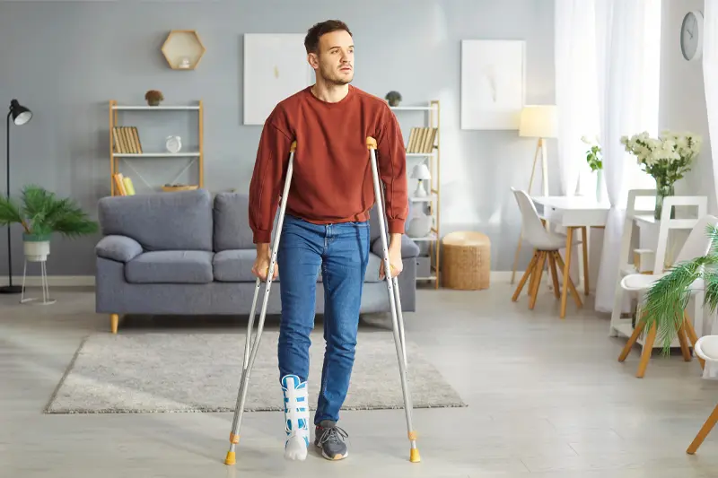 A male client stands in a therapy room on crutches with one leg in a boot