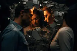 A man and woman face each other, faces framed by broken shards, while a third figure of a woman stands beyond them in the foreground