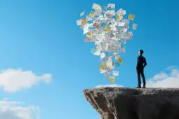 A male figure looks upward at a cloud of paperwork while standing near the edge of a cliff
