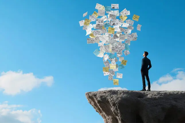 A male figure looks upward at a cloud of paperwork while standing near the edge of a cliff