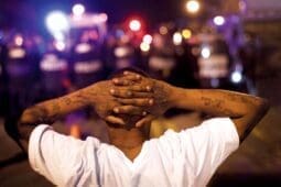 A Black man faces the police and holds his hands behind his head