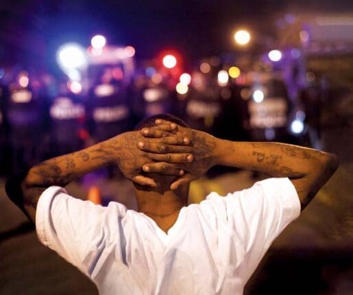 A Black man faces the police and holds his hands behind his head