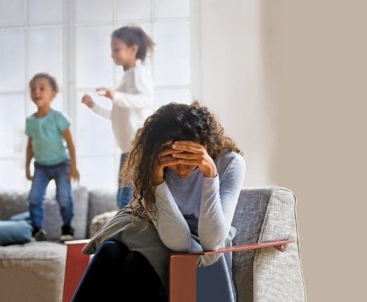 A mother holds her head in her hands as her children jump on the couch behind her