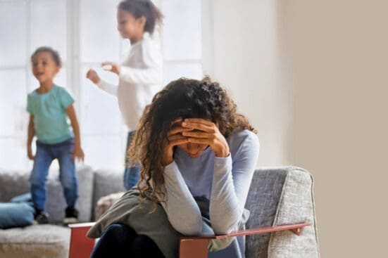 A mother holds her head in her hands as her children jump on the couch behind her