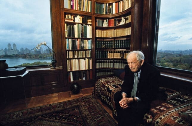 A man sits next to a full bookshelf