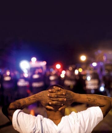 A Black man faces the police and holds his hands behind his head