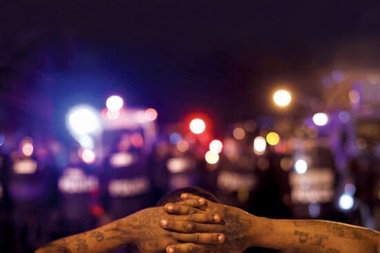 A Black man faces the police and holds his hands behind his head