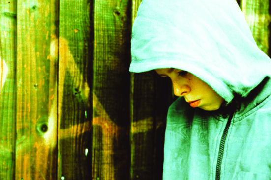 A young boy with a hood on stands next to a fence and looks dejected