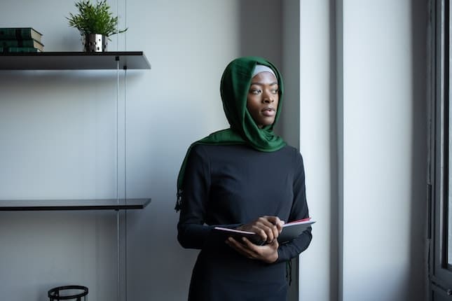 Black woman with a head scarf stares out the window - Photo Credit: Pexels/Monstera