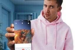 A man in a pink hoodie holds a phone toward the camera's view, showing a dating app profile